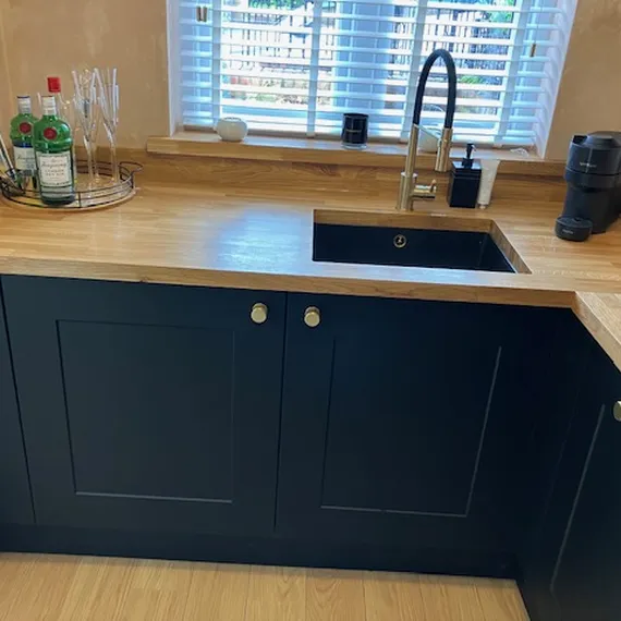 Modern kitchen corner with black cabinets, wooden countertop, sink with faucet, liquor bottles, glasses, coffee maker, and window with blinds.