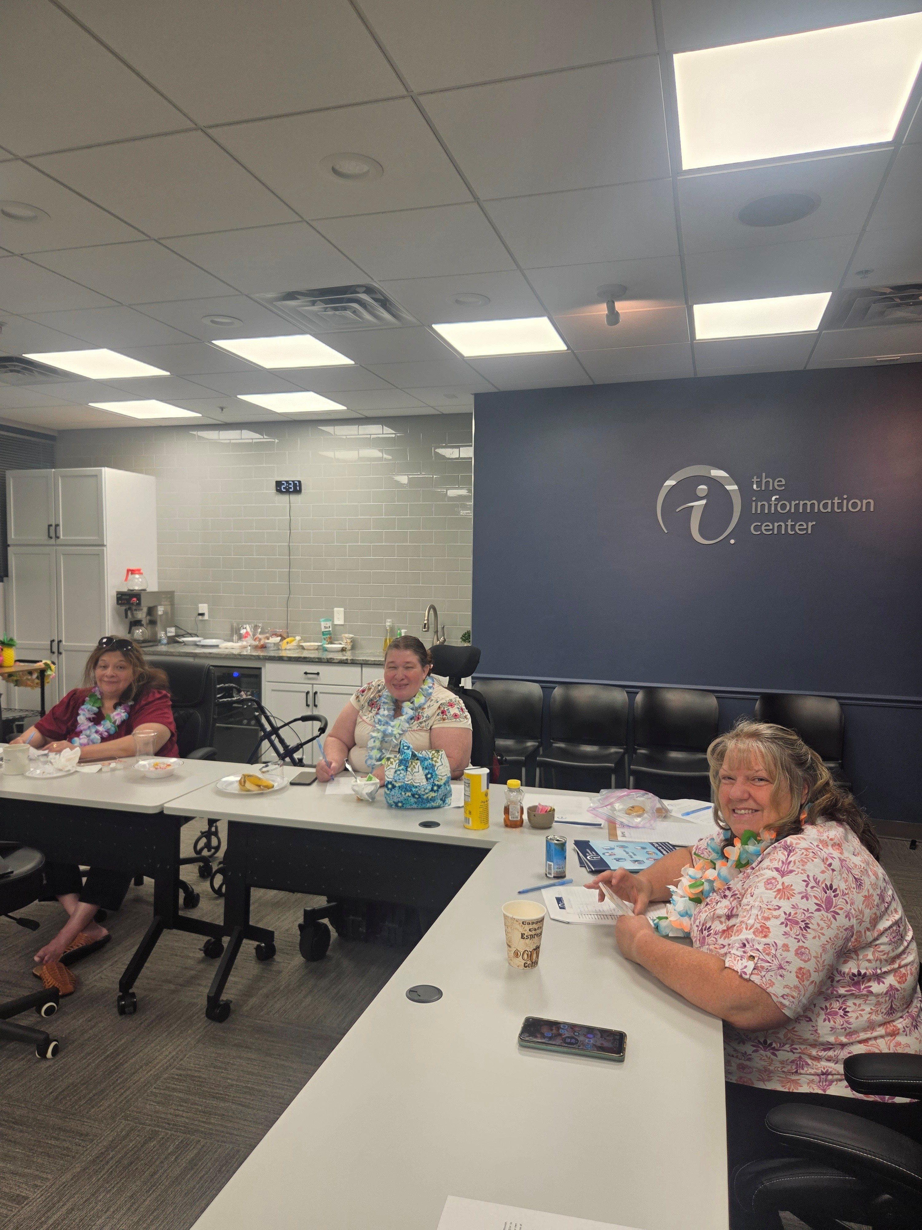 Three women sitting at a conference room table with leis, smiling, with snacks and drinks in front of them. A sign behind them reads "the information center."