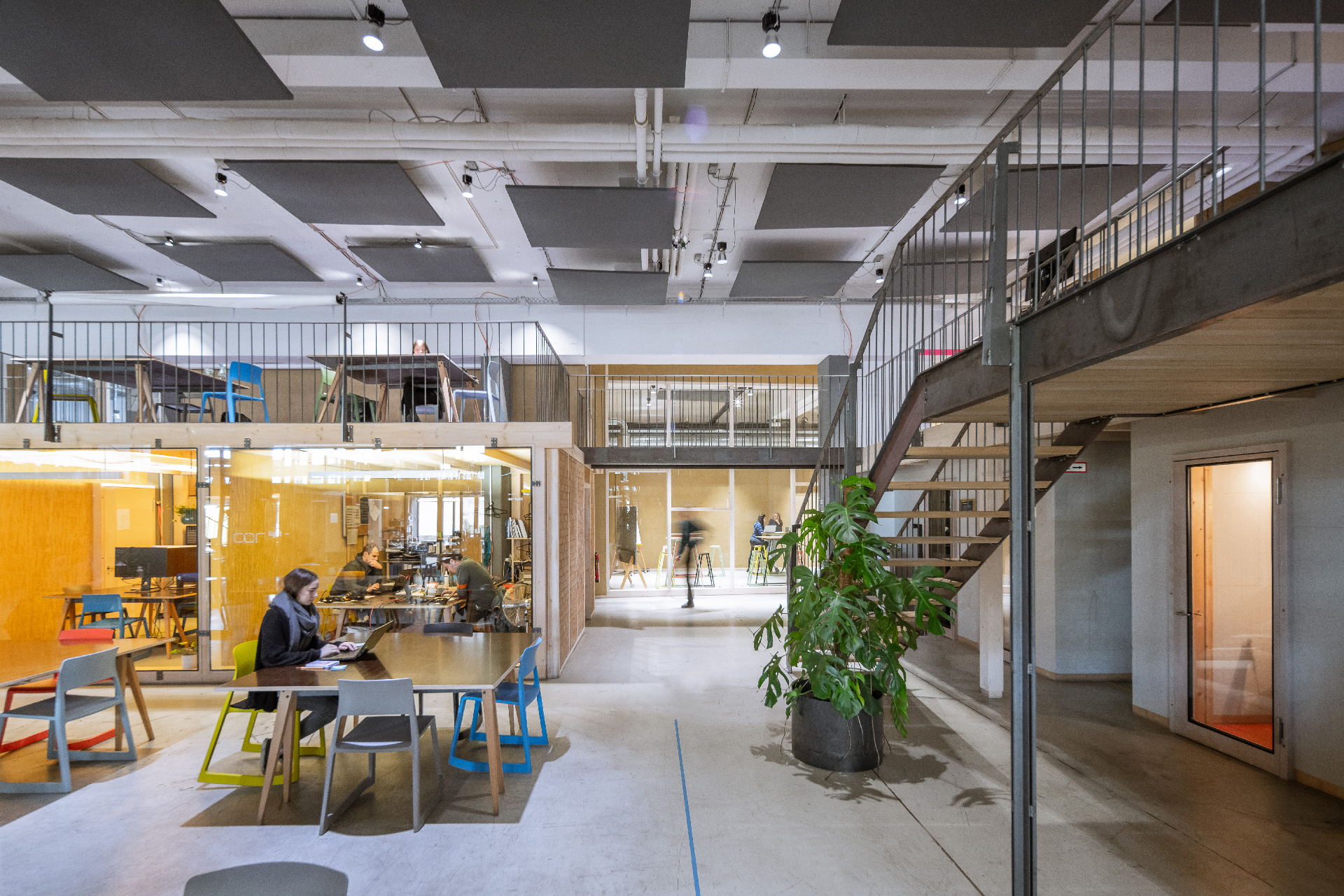 Heller, moderner Coworking-Space mit Glasbüros, Galerieebene und arbeitenden Personen an Tischen in einem offenen Raum.
