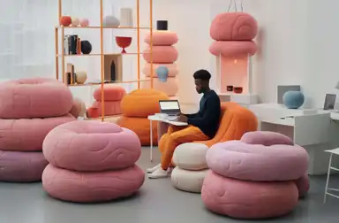 A modern, colorful living space featuring a person sitting on an orange chair and working on a laptop, surrounded by unique, rounded pink furniture pieces.