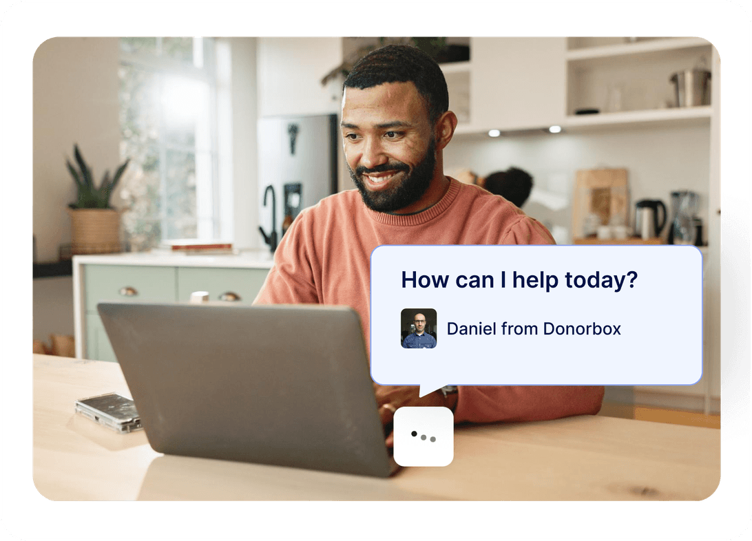 Man smiling at a laptop in a kitchen, chat bubble displaying "How can I help today? Daniel from Donorbox."