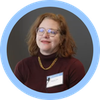 Person with red curly hair and glasses wearing a dark maroon top with a gold necklace and a name badge, circular photo.