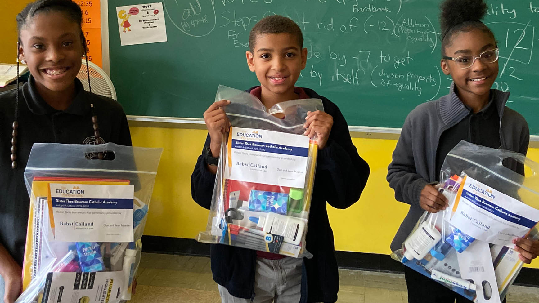 Three smiling students holding school supply kits from Sister Thea Bowman Catholic Academy's Adopt-A-School program in a classroom.