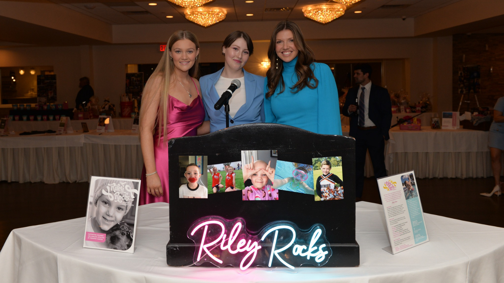 Three women posing at a Riley Rocks Foundation charity event display with neon sign and children's photos in a banquet hall.