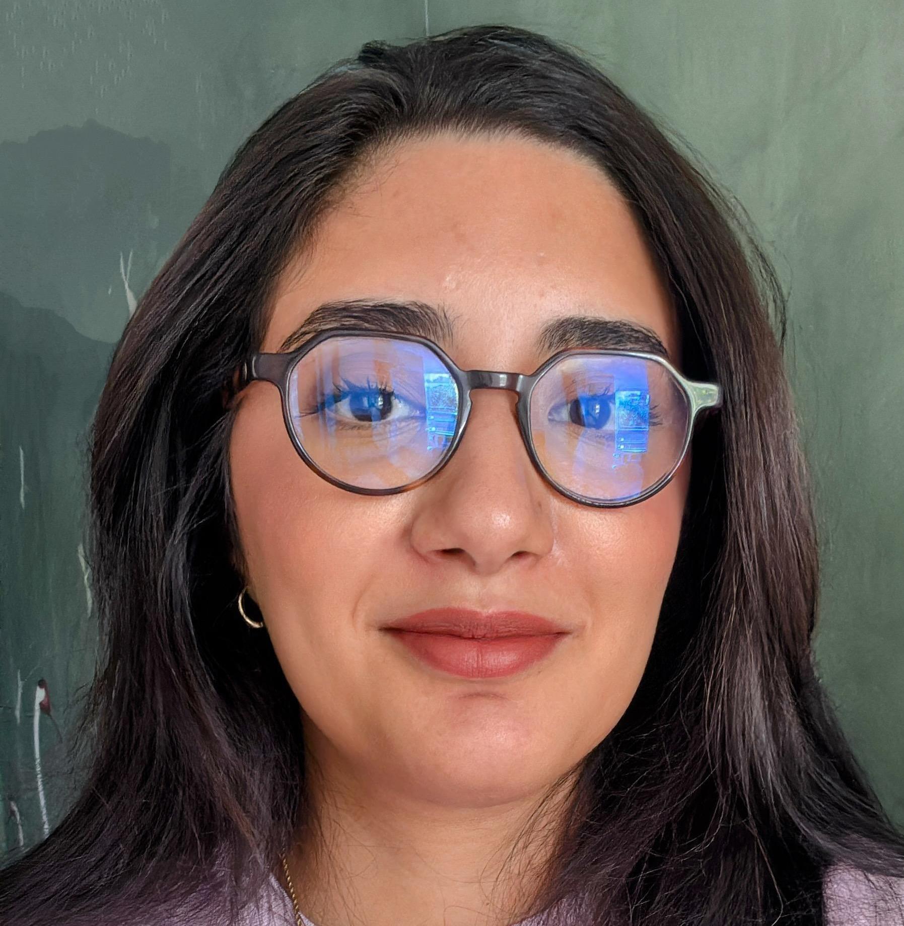 Woman with long dark hair wearing geometric tortoiseshell glasses with blue light reflection, subtle smile, against a green background.