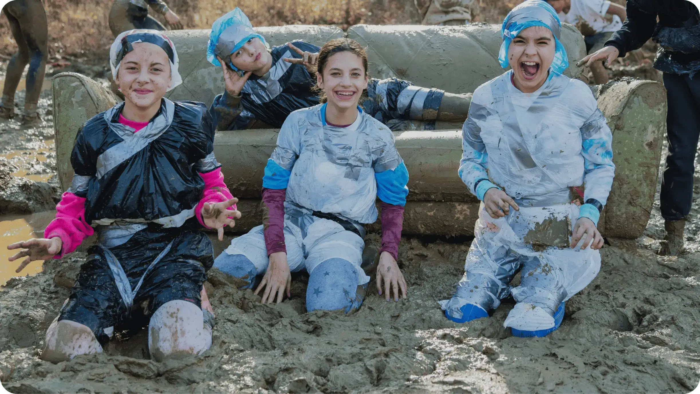 Three muddy children laughing and posing during an outdoor mud run obstacle course event, wearing protective gear and shower caps.