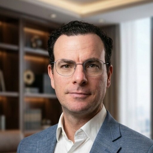 Professional man with glasses and curly dark hair wearing a gray blazer, posed in a modern office with bookshelves in background.