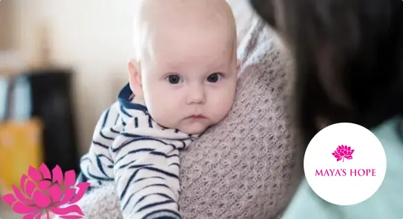 Baby in striped outfit lying on a person's shoulder, looking intently at the camera. Maya’s Hope logo in the corner.