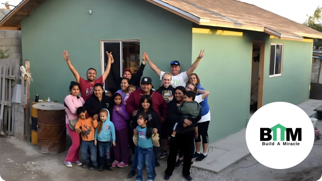 A group of people joyfully pose in front of a small, newly built green house. The Build a Miracle logo is visible in the corner.