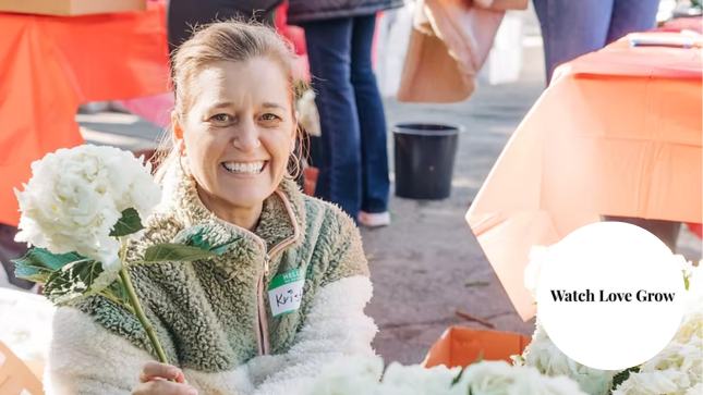 Woman smiling while holding white flowers at an outdoor event with orange tables; text reads "Watch Love Grow."