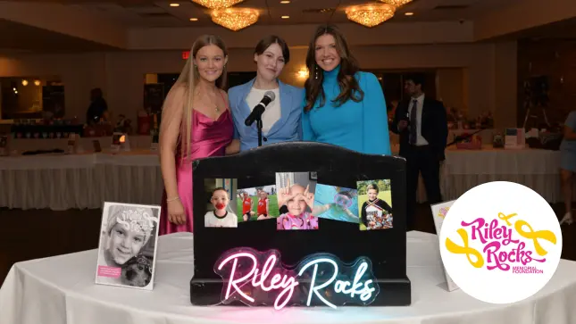 Three people stand behind a display with "Riley Rocks" in neon. The table features photos and a poster. The room has elegant lighting.