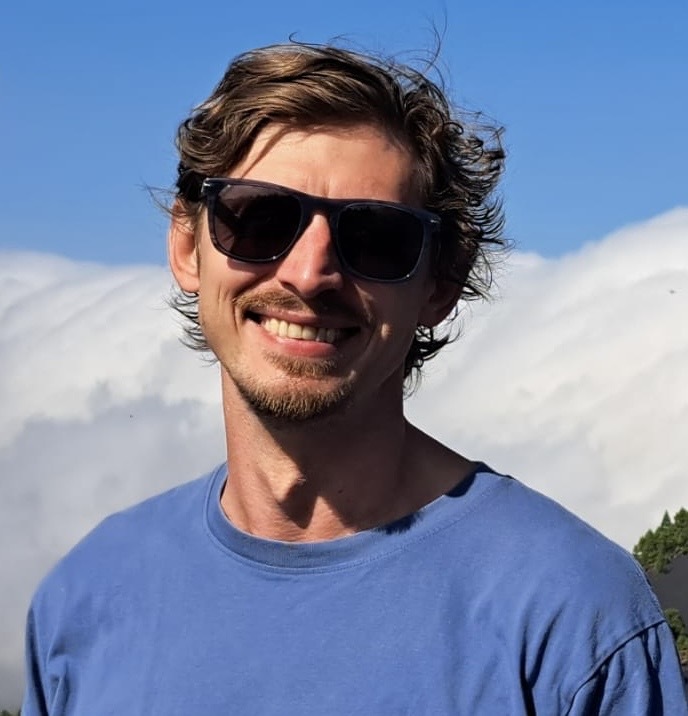 Smiling young man with curly brown hair wearing sunglasses and a blue t-shirt, with a bright blue sky and clouds behind him.