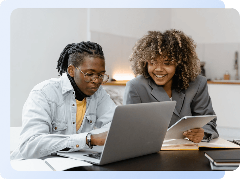 Two people, one looking at a laptop and the other holding a tablet, smiling and collaborating at a desk.