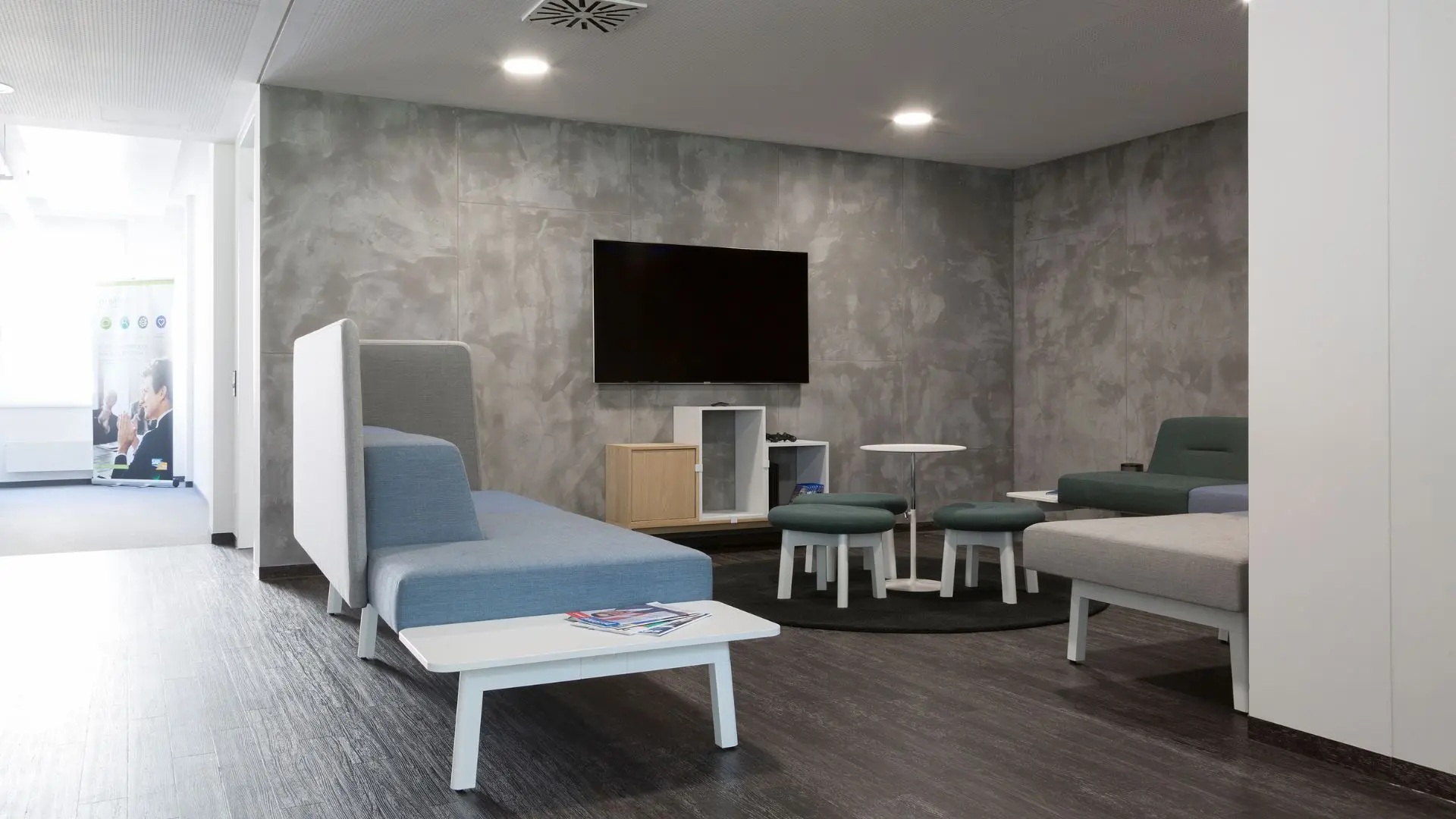 Modern office lounge with gray couches, flat-screen TV on a textured wall, round tables, blue stools, and magazines on a low table.
