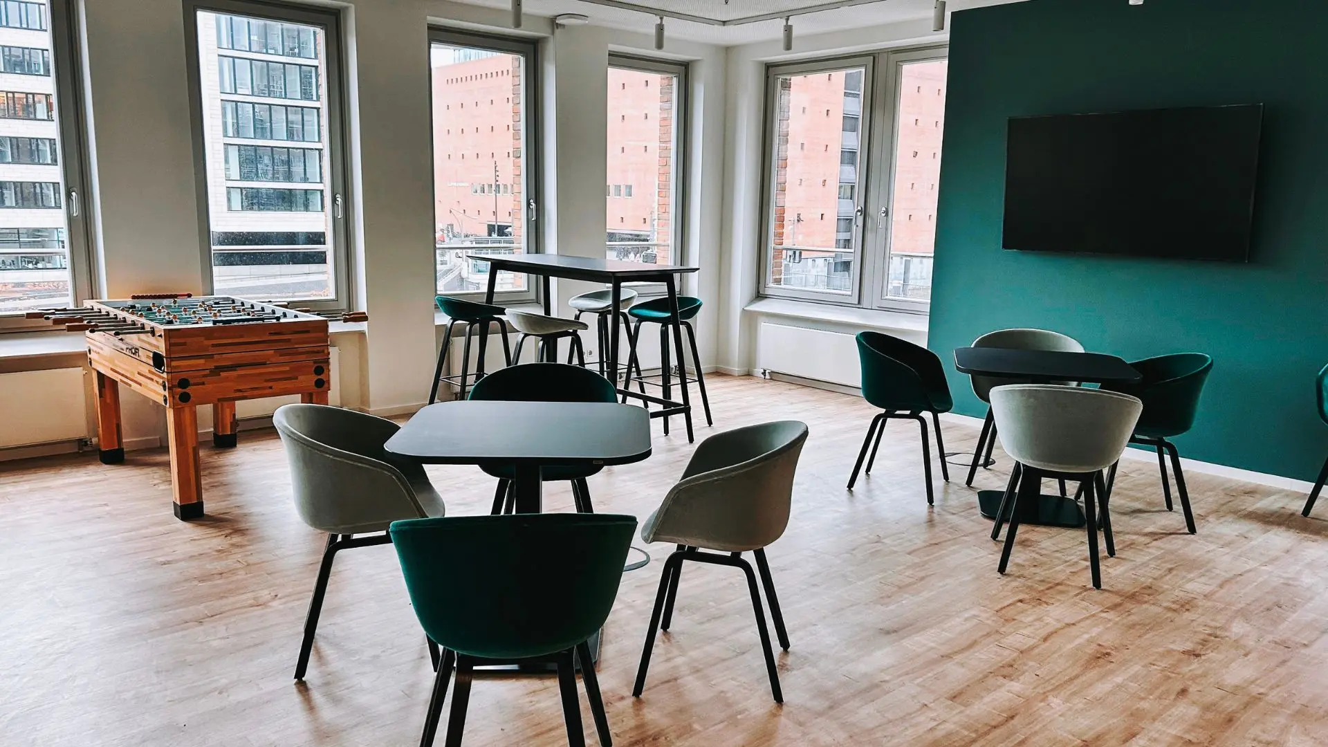 Modern lounge with large windows, tables, chairs, a high table, and a foosball table. Bright, spacious, and with wooden flooring.