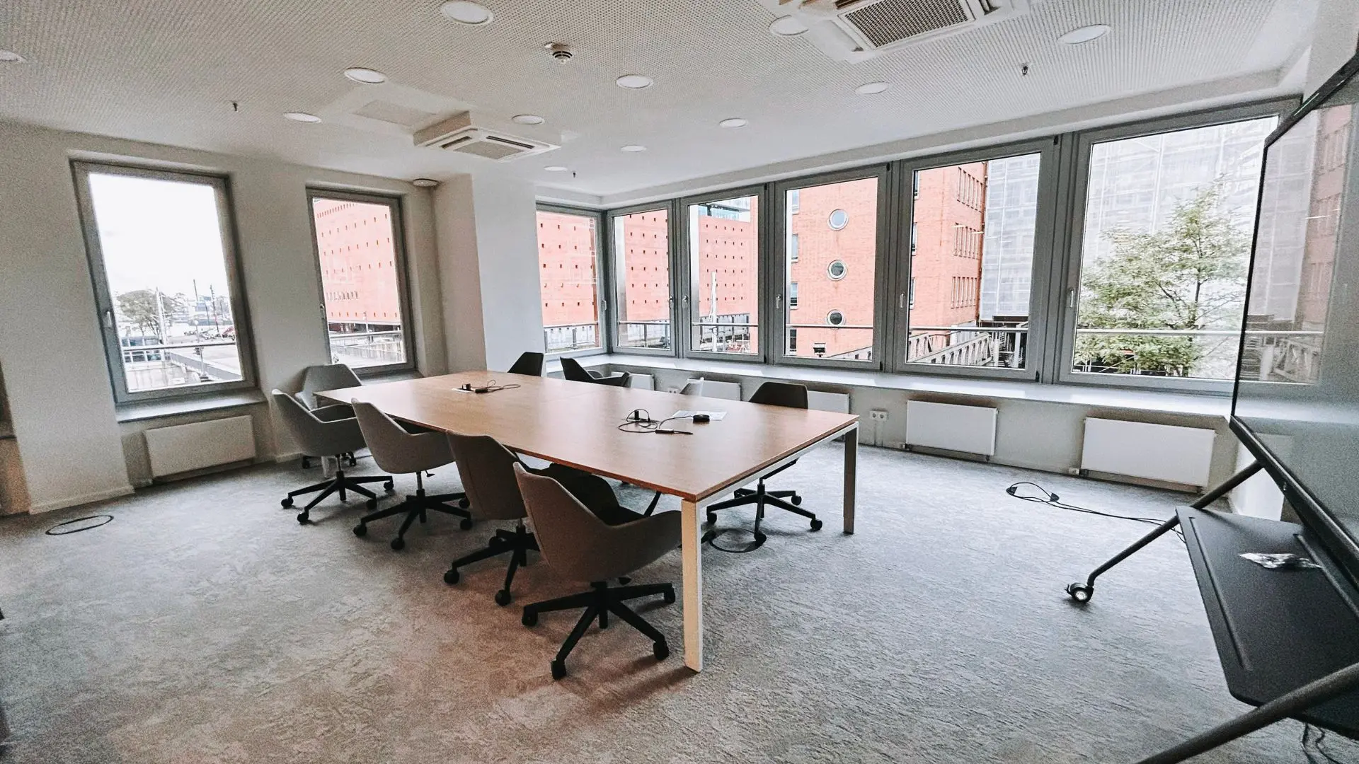 Modern conference room with a wooden table, swivel chairs, large windows, and glass writing board, offering a city view.