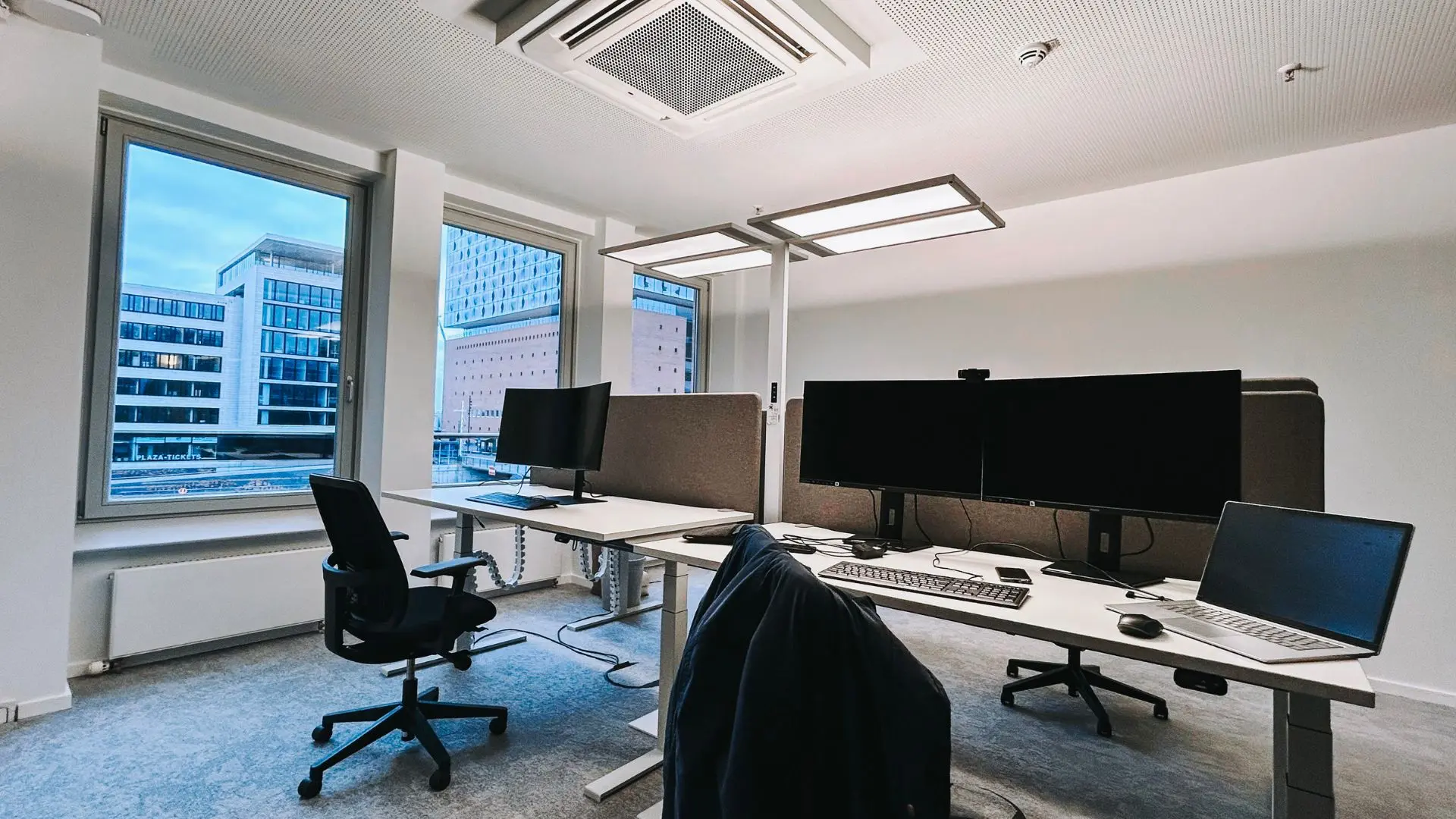Modern office space with desks, ergonomic chairs, computers, and large windows offering a city view. Minimalist design with neutral colors.