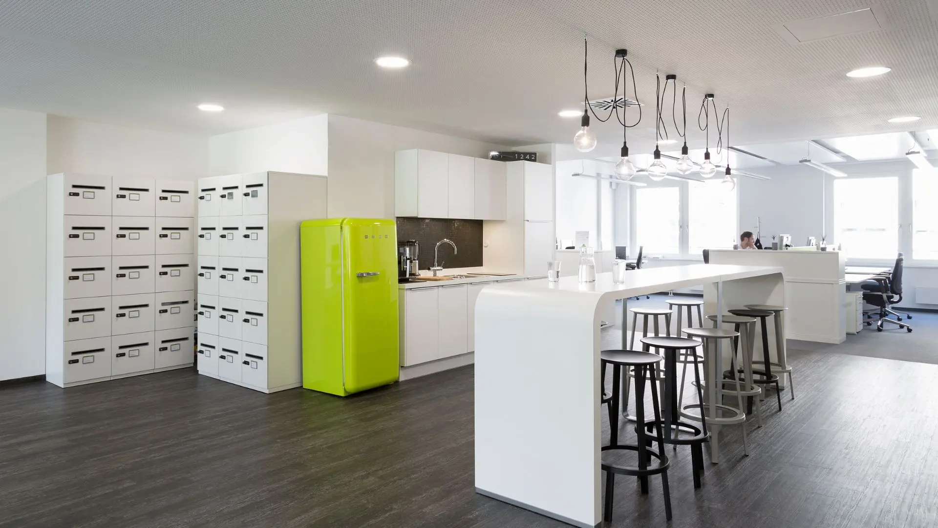 Modern office kitchen with white cabinets, a bright green fridge, high tables with stools, and multiple pendant lights on a dark wood floor.