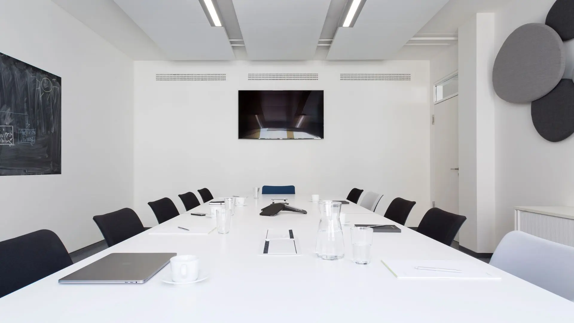 A modern conference room with a long white table, black and white chairs, a wall-mounted TV, a laptop, water glasses, and notepads.