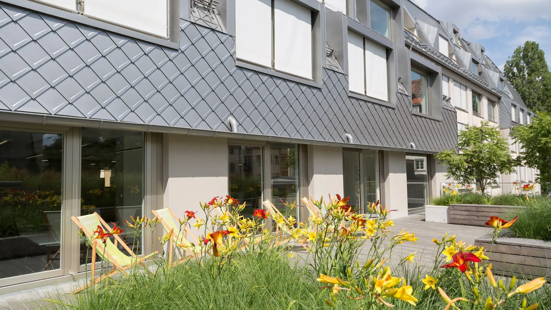 Roof terrace with planted yellow and red flowers in the foreground, and deck chairs on a patio.