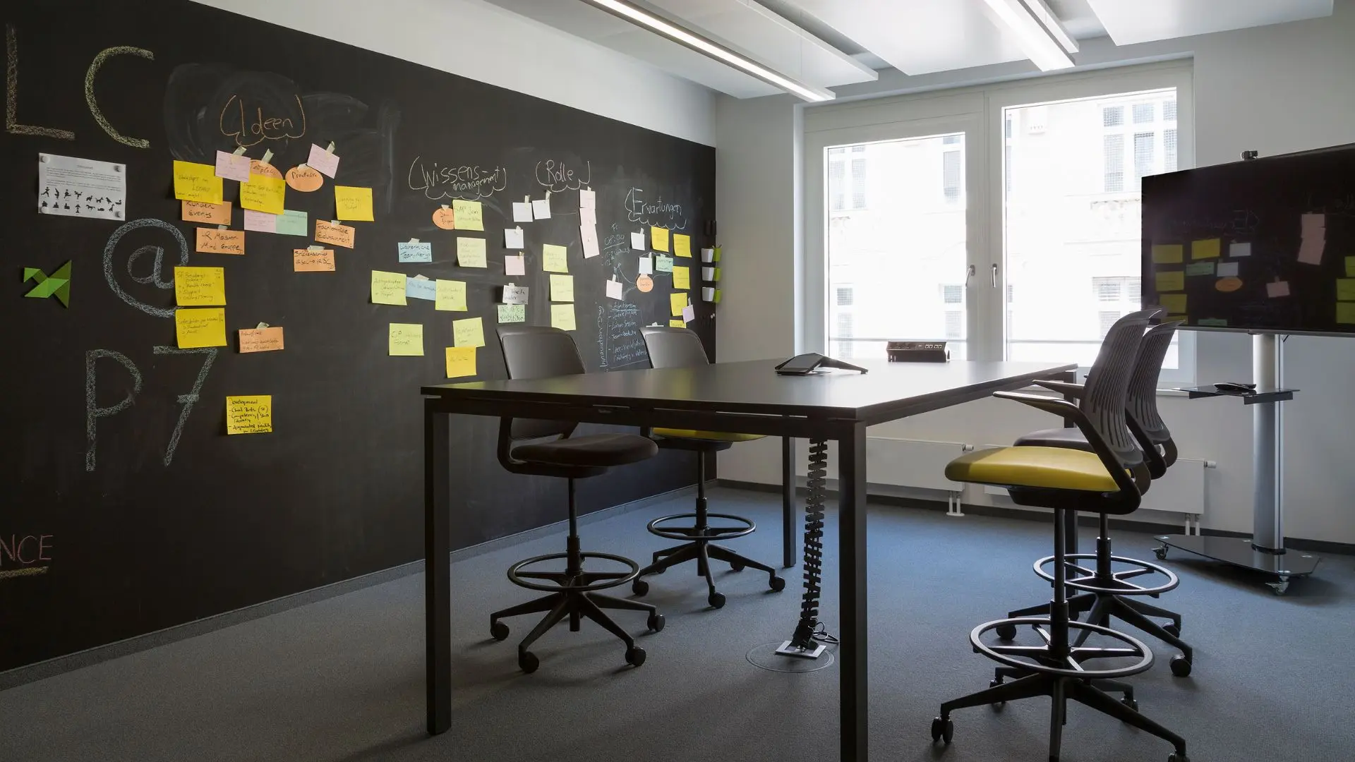 Modern office with a large blackboard wall covered in colorful sticky notes, a high table with chairs, and a mounted TV.