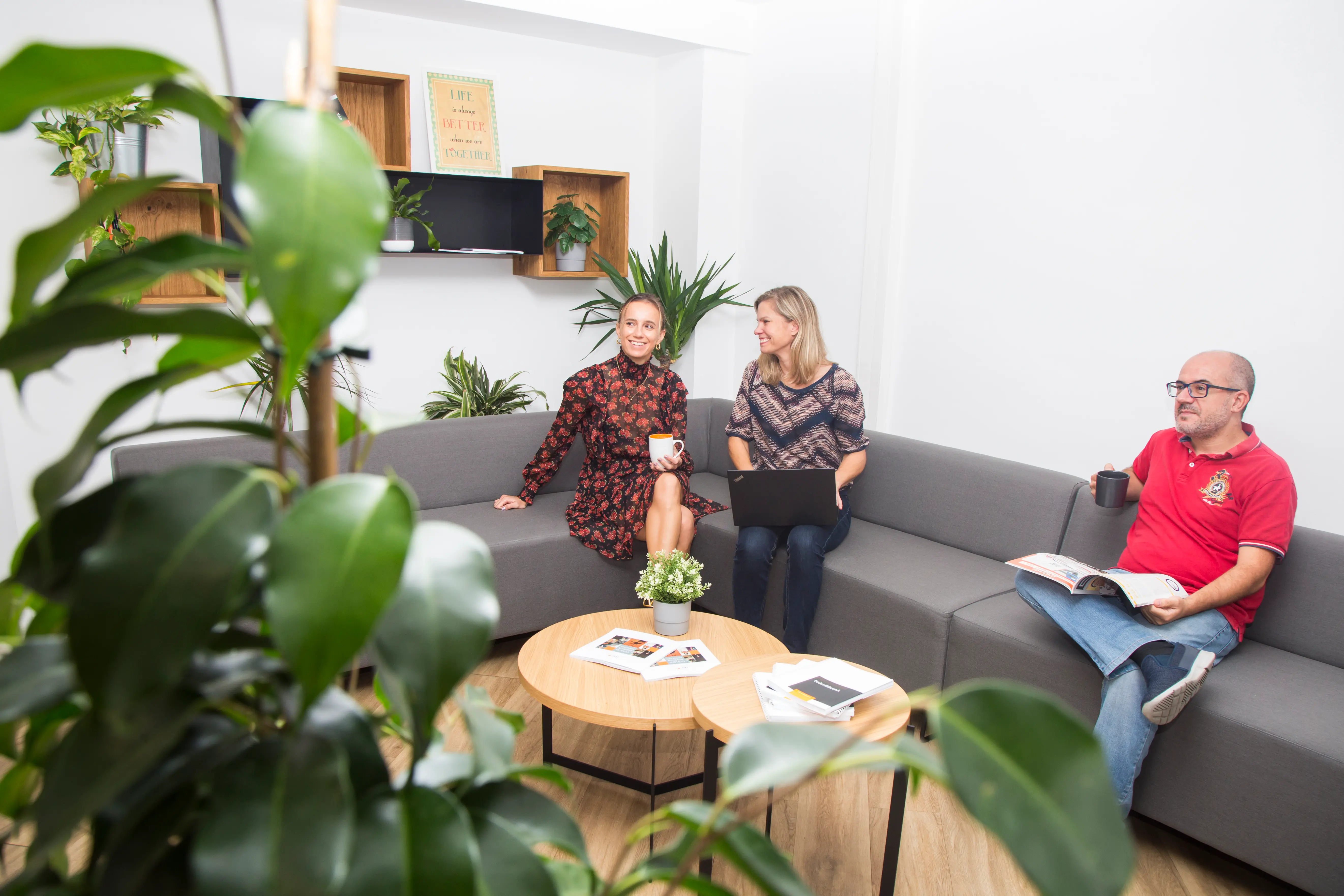 Three people in a modern, plant-filled office lounge area, sitting on a gray couch with coffee, a magazine, and a laptop.