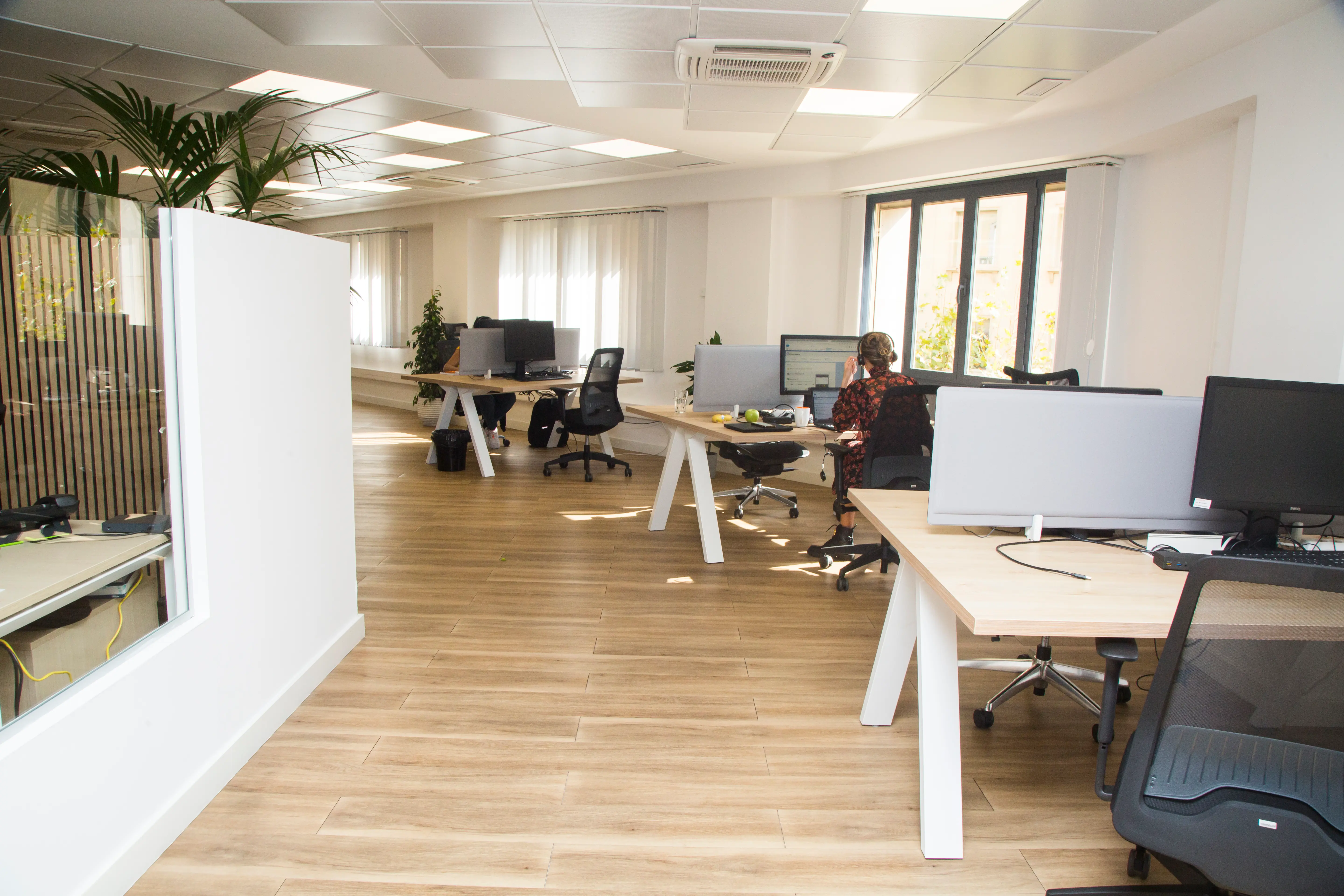 Modern open-plan office with wooden flooring, plants, and white walls. Desks have computers and chairs. A person works at a desk near the window.
