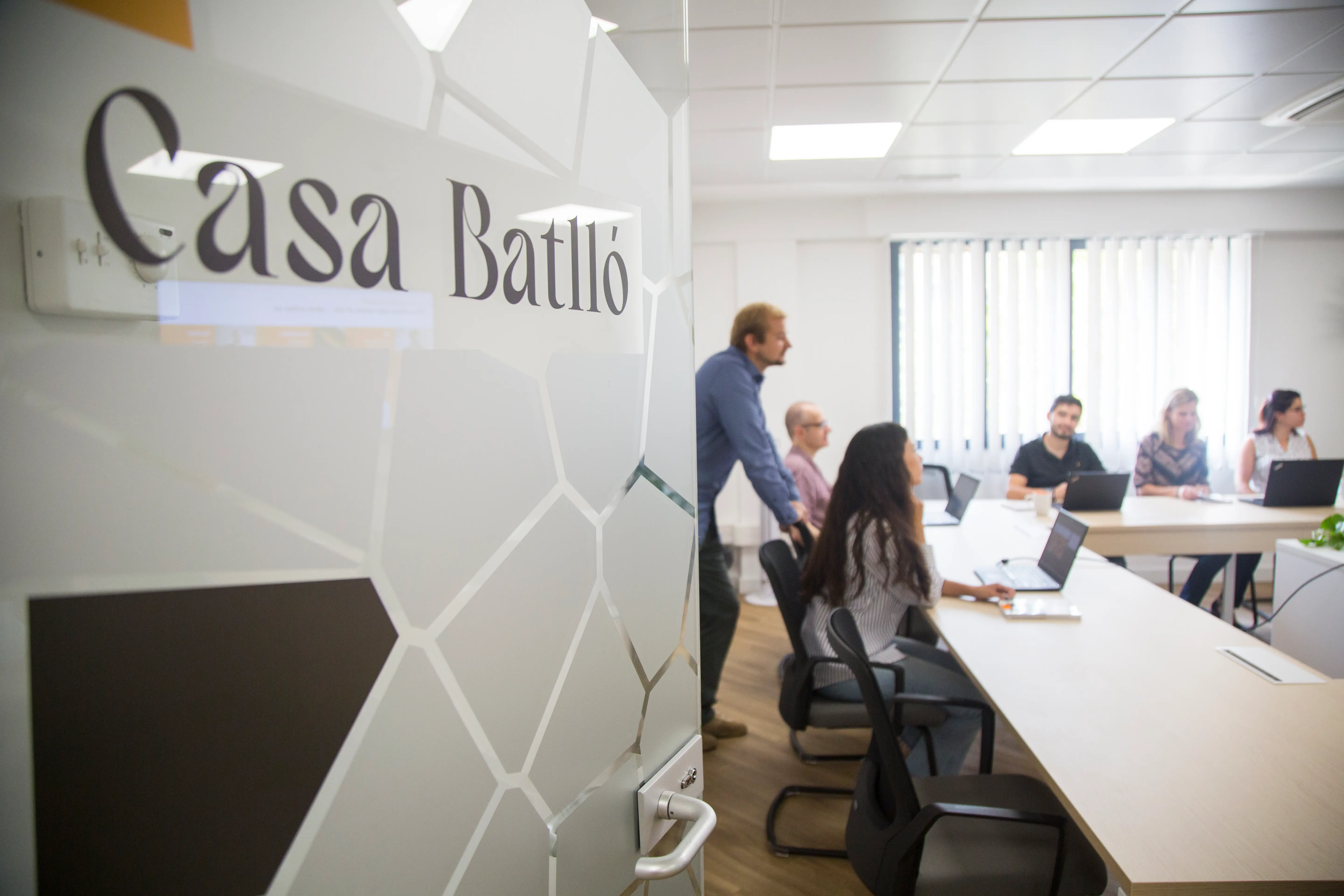 Office meeting with people around a table and laptops, viewed through a glass door labeled "Casa Batlló."