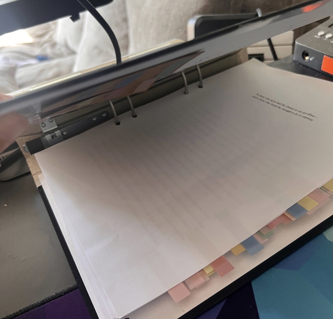 An open binder with a stack of papers and colored sticky tabs, viewed from the side on a desk, with a laptop partially visible above.