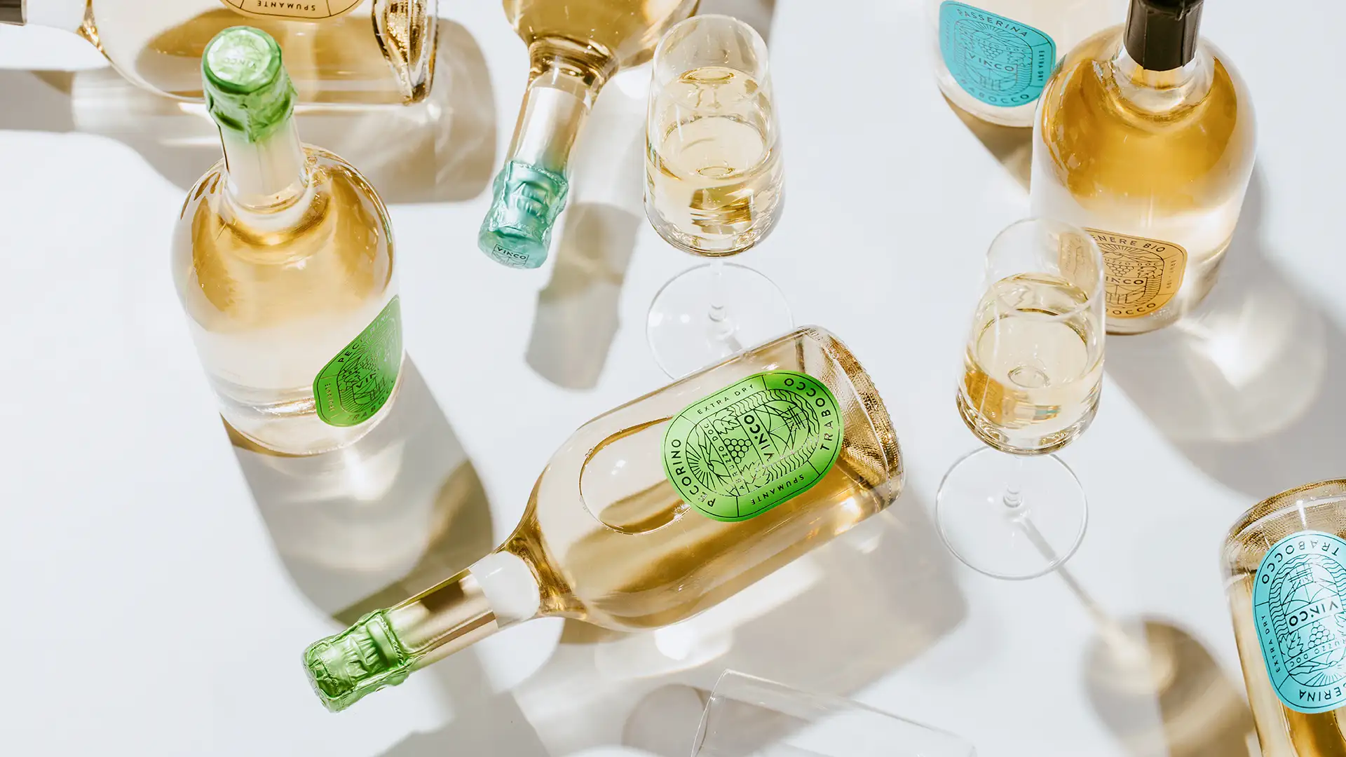 Vista dall'alto di bottiglie e calici di Vinco Spumante Pecorino su sfondo bianco, con spumante versato nei bicchieri