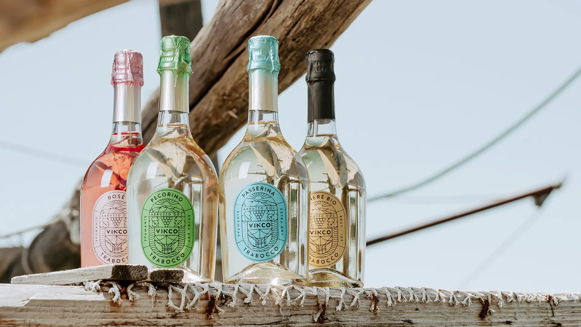 Quattro bottiglie di Vinco Spumante (Pecorino, Passerina, Rosé e Venere Bio) appoggiate ai pali di un trabocco con il mare sullo sfondo