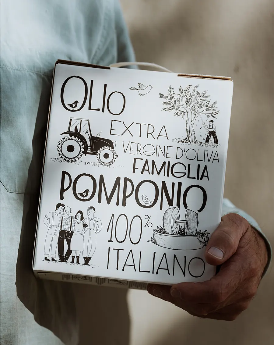 Dettaglio della bottiglia Famiglia Pomponio con tappo nero e packaging illustrato, su superficie in legno con vegetazione sfocata sullo sfondo