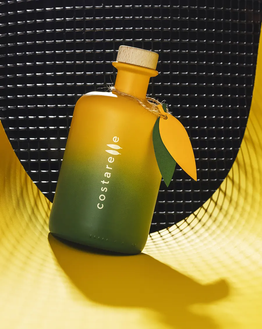 Bottiglia Costarelle con packaging verde sfumato e pendaglio in legno a forma di foglia, appoggiata su cestino intrecciato con sfondo a griglia scura