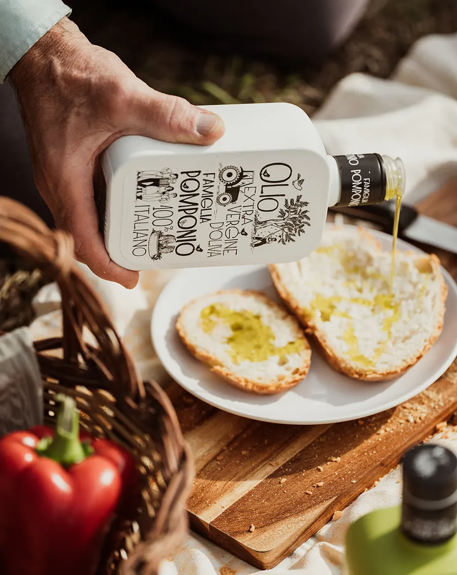 Mano che versa Olio Extravergine Famiglia Pomponio su fette di pane tostate, con verdure e cesto sullo sfondo