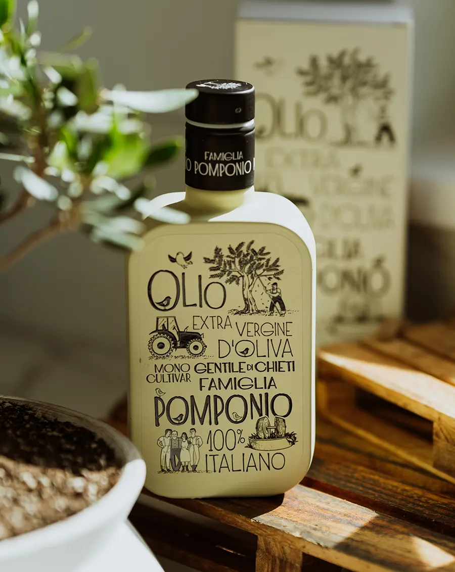 Bottiglia di Olio Extravergine Monocultivar Gentile di Chieti Famiglia Pomponio su tagliere in legno con sfondo di packaging e piante d'ulivo