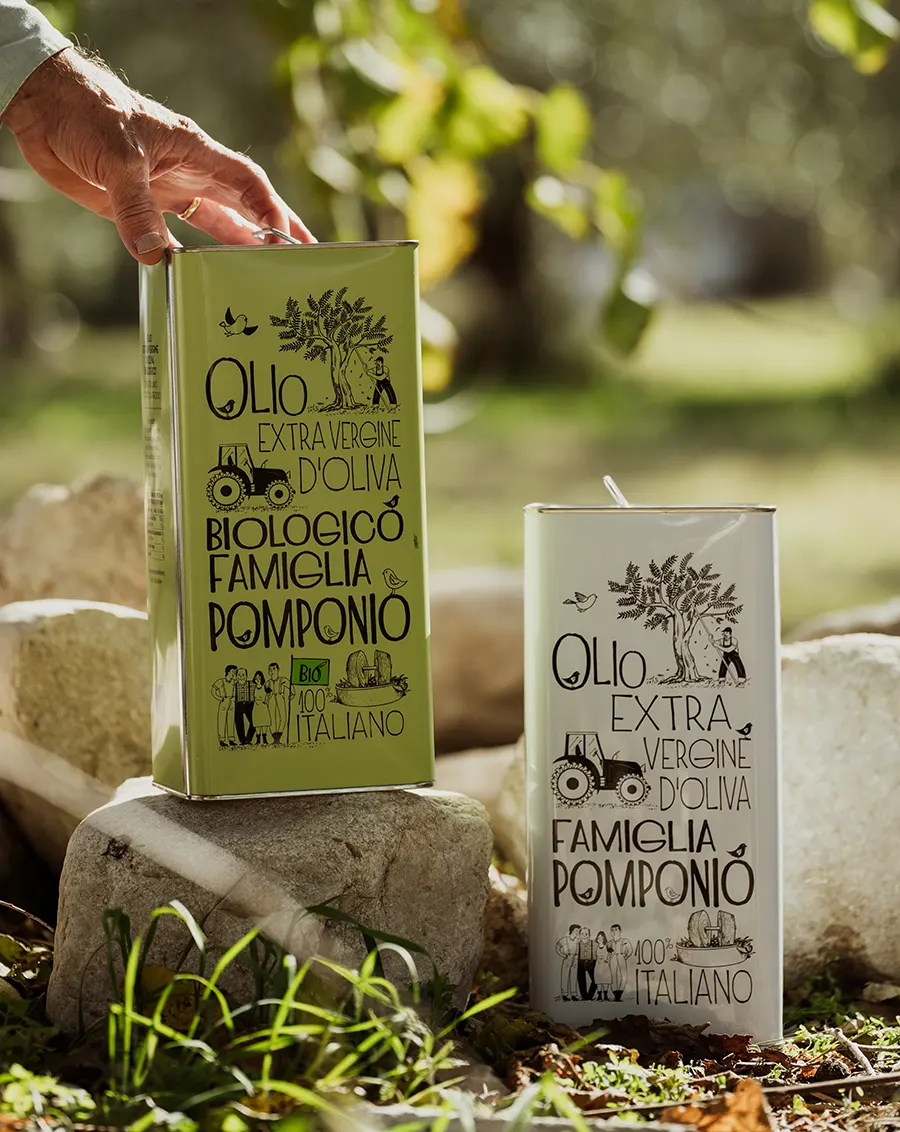 Mano che afferra una lattina di Olio Extravergine Biologico Famiglia Pomponio accanto a una piccola latta classica, in un contesto naturale con erbe