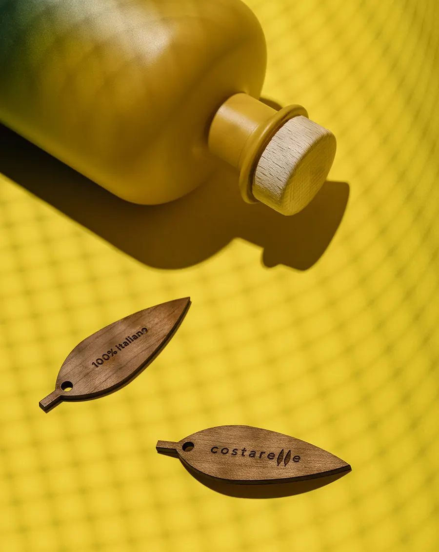 Primo piano del tappo in legno e del pendaglio in legno inciso con il logo Costarelle e la scritta "100% italiano", su sfondo a rete gialla