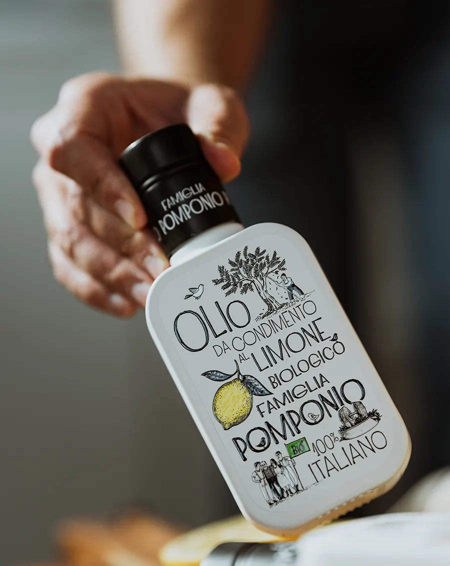 Mano che inclina la bottiglia Famiglia Pomponio con etichetta "Olio da Condimento al Limone Biologico" su sfondo scuro sfocato
