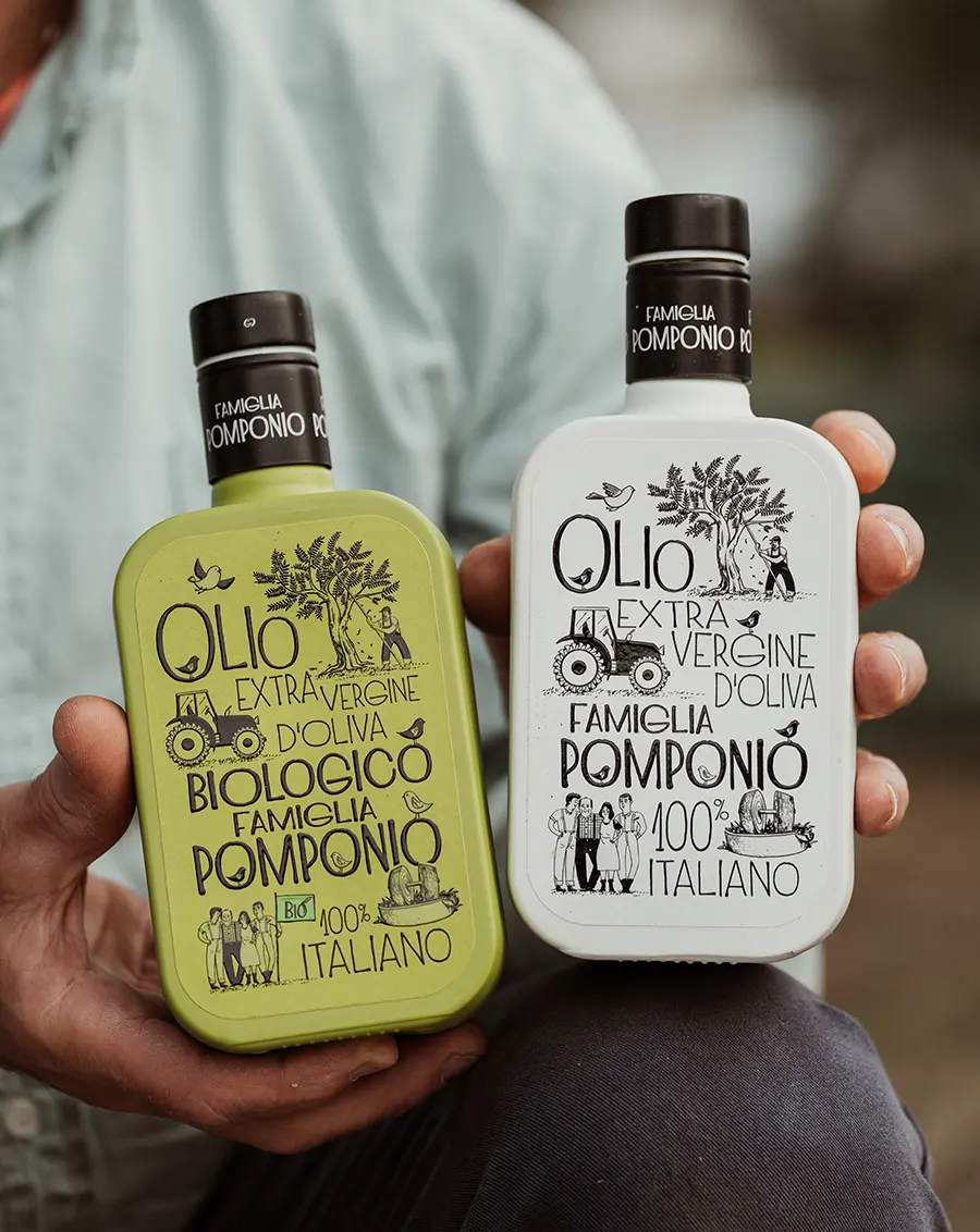 Mano che regge due bottiglie di Olio Famiglia Pomponio, versione classica e biologica, con etichette illustrate in stile artigianale