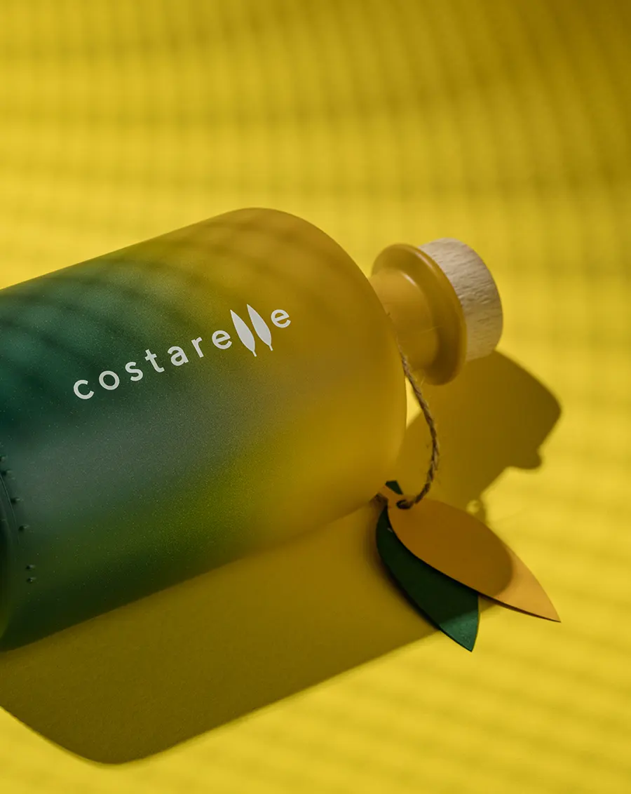 Dettaglio ravvicinato della bottiglia Costarelle verde con logo bianco in rilievo e pendaglio a foglia, su sfondo giallo caldo
