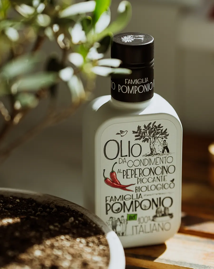 Bottiglia di Olio da Condimento al Peperoncino Piccante Biologico Famiglia Pomponio tra le foglie di un ulivo in piena luce naturale