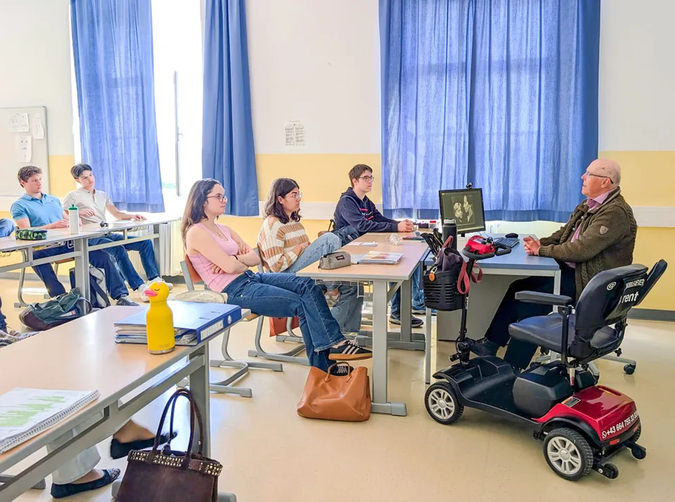 Alter Mann an einem Lehrertisch sitzend auf in Richtung Schüler:innen sprechend.