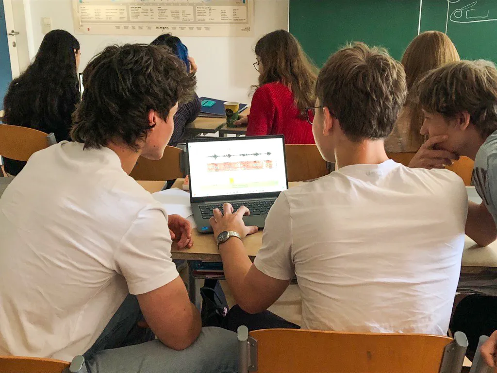 Drei männliche Jugendliche in einem Klassenzimmer vor einem Laptop sitzend auf den Bildschirm blickend mit dem Rücken zur Kamera