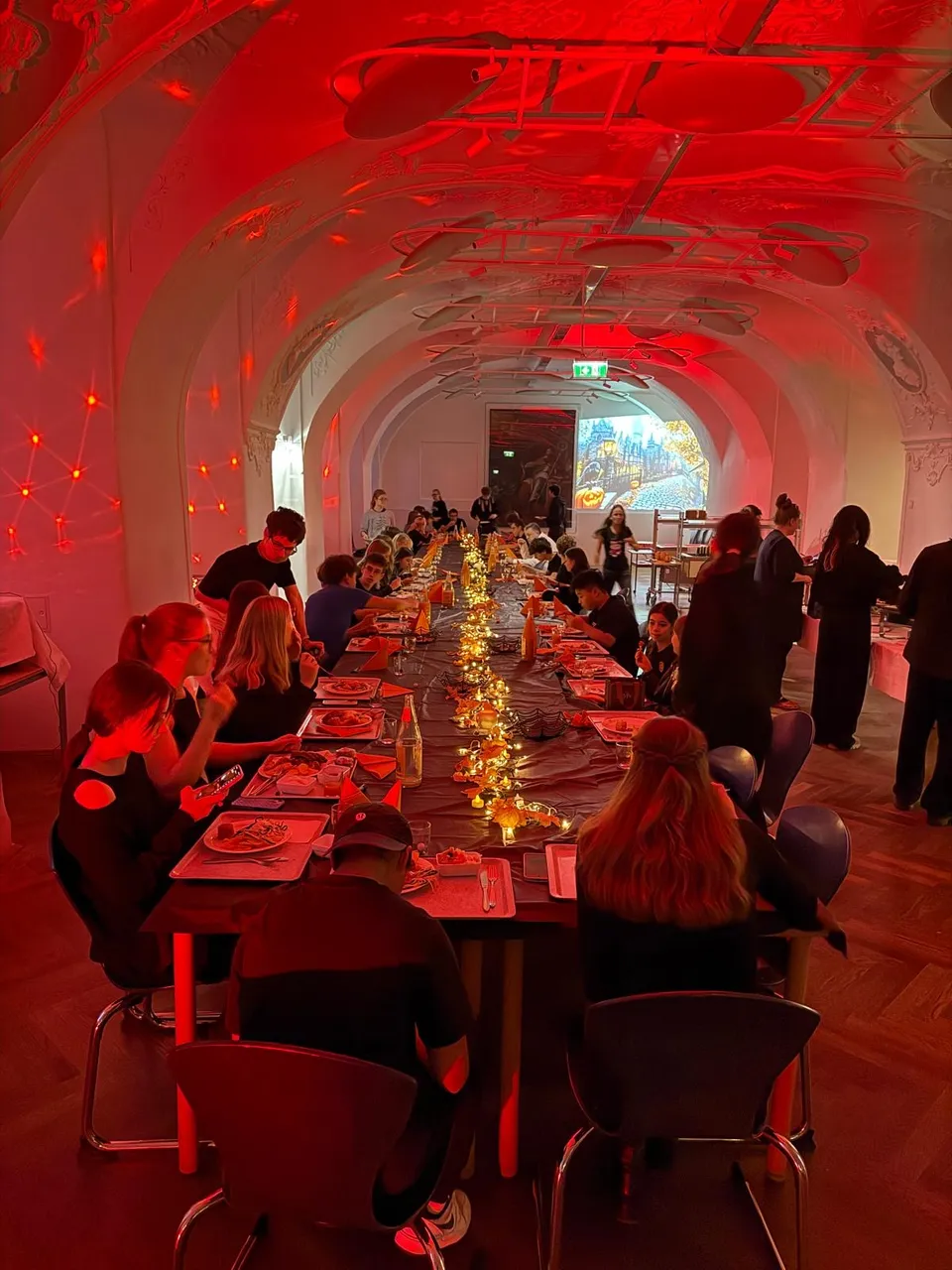 A long dining table in a red-lit room, set with plates and candles, with people seated and standing around, creating a warm, festive atmosphere.