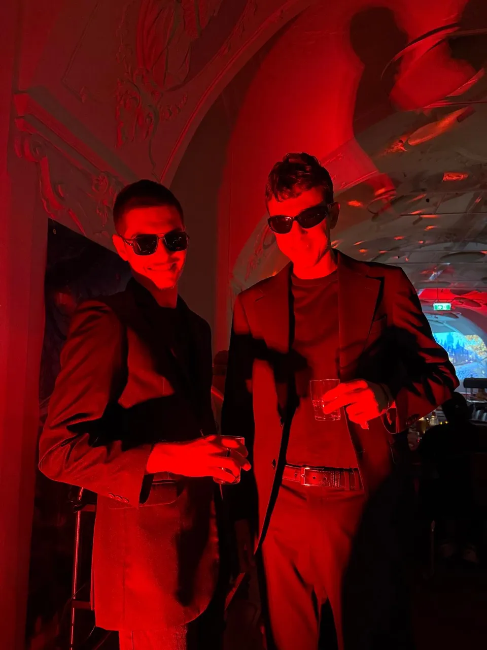 Two people wearing sunglasses and dark suits stand in a dimly lit room with red lighting, holding drinks and smiling.