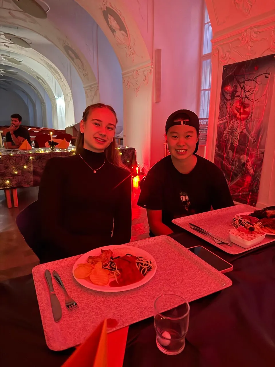 Two people sitting at a table in a red-lit room with ornate arches, dining on plates of food including pasta, chips, and salad.