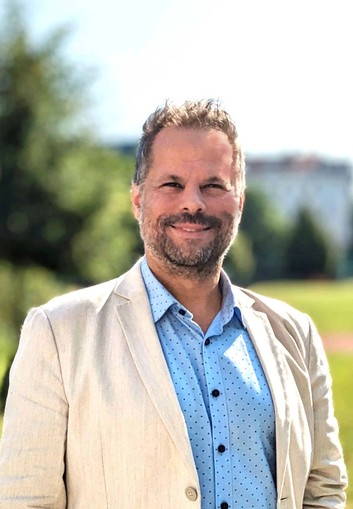 Portrait eines Mannes mit leicht ergrautem Vollbart, blauen Hemd und hellem Sakko freundlich in die Kamera lachend vor einem verschwommenem parkähnlichem Hintergrund