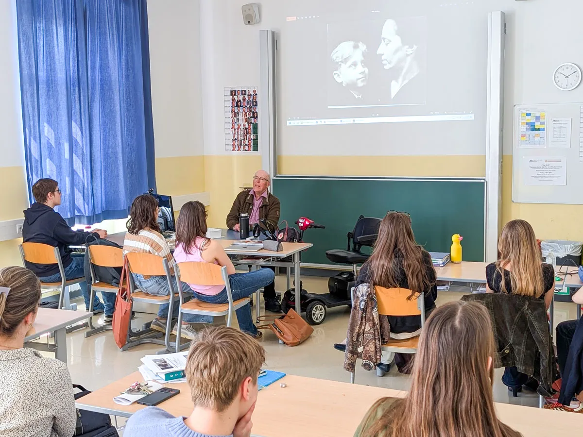 Alter Mann mit Brille in einem Klassenzimmer am Lehrertisch sitzend und zu Jugendlichen sprechend.