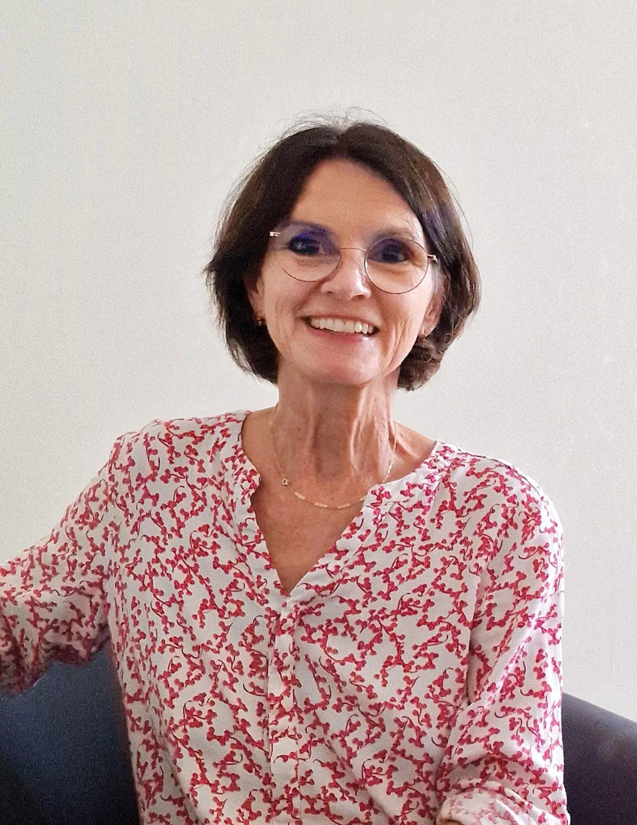 sympathisch lachende reifere Frau mit Pagenschnittfrisur, großer Brille und rot weiß gemusterter Bluse in einem Stuhl sitzend vor einem neutralen Hintergrund