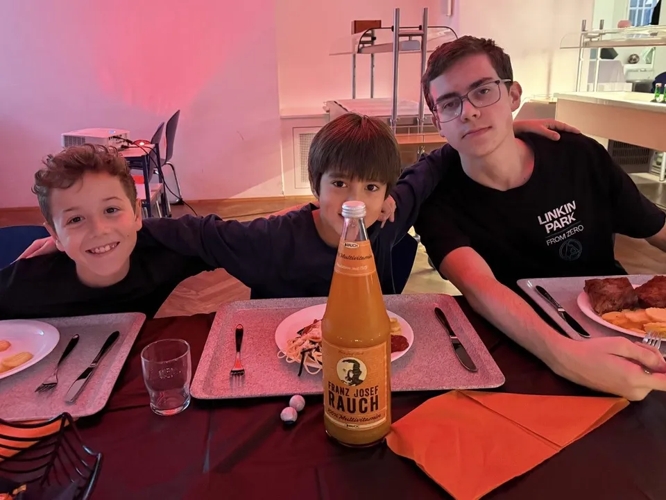 Three boys smiling and sitting at a table with food and drinks, including a large bottle labeled "Rauch," in a warmly lit room.